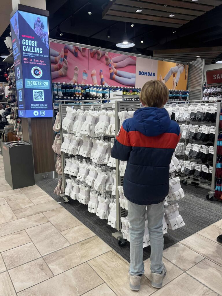 A person in a blue and red jacket stands in a store aisle looking at a display of white socks. There is a digital sign on the wall promoting an event called "Goose Calling" with ticket information. The store has tiled floors and overhead lighting.
