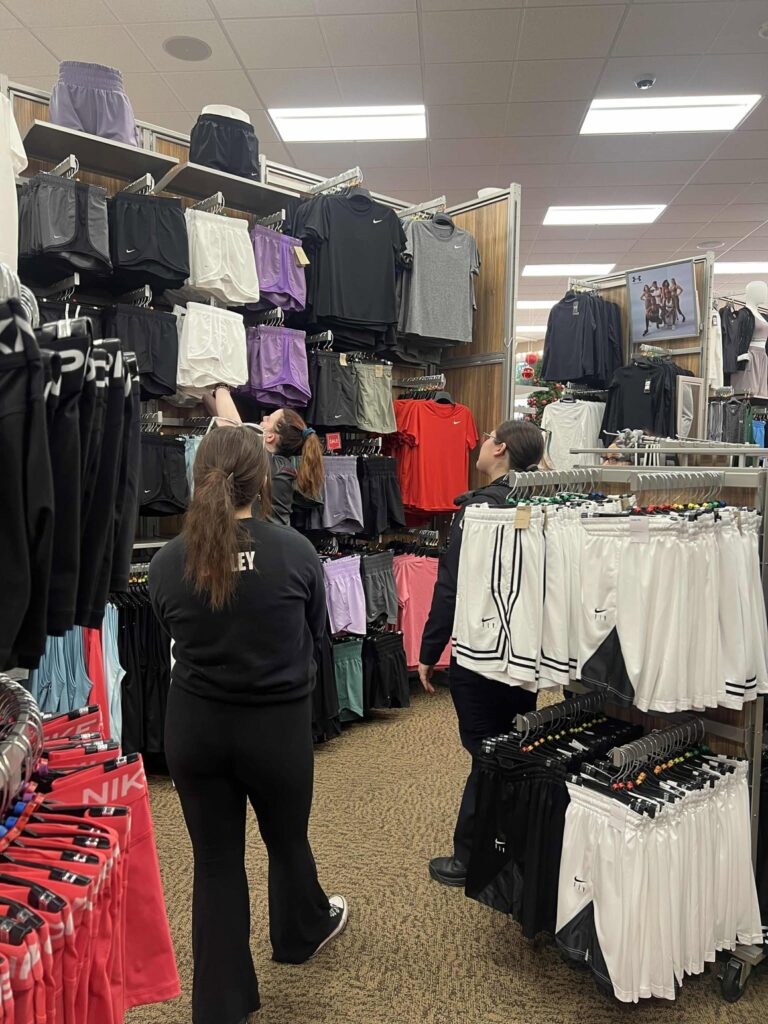 Three people browse athletic clothing in a retail store. Clothes are neatly displayed on racks and shelves, including shorts, shirts, and pants in various colors. The store has bright lighting and a carpeted floor.