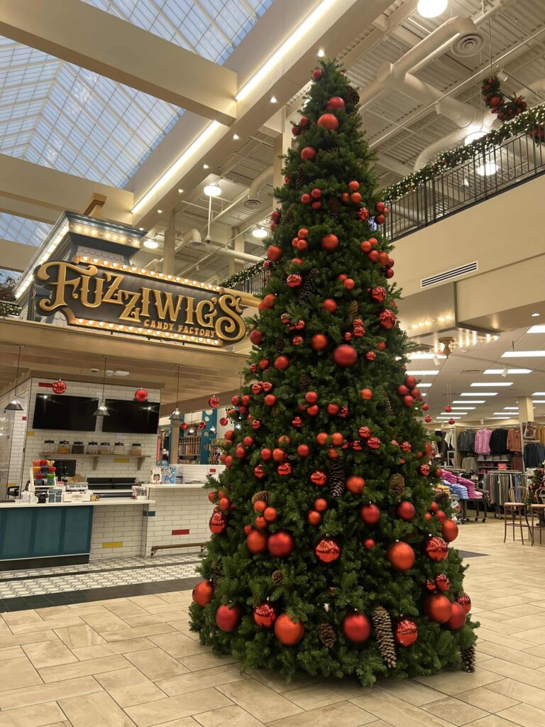 A tall Christmas tree decorated with red ornaments and pinecones stands in a shopping mall. Nearby, a store sign reads "Fuzziwig's Candy Factory." The ceiling features a grid design with large windows. Clothing displays are visible to the right.