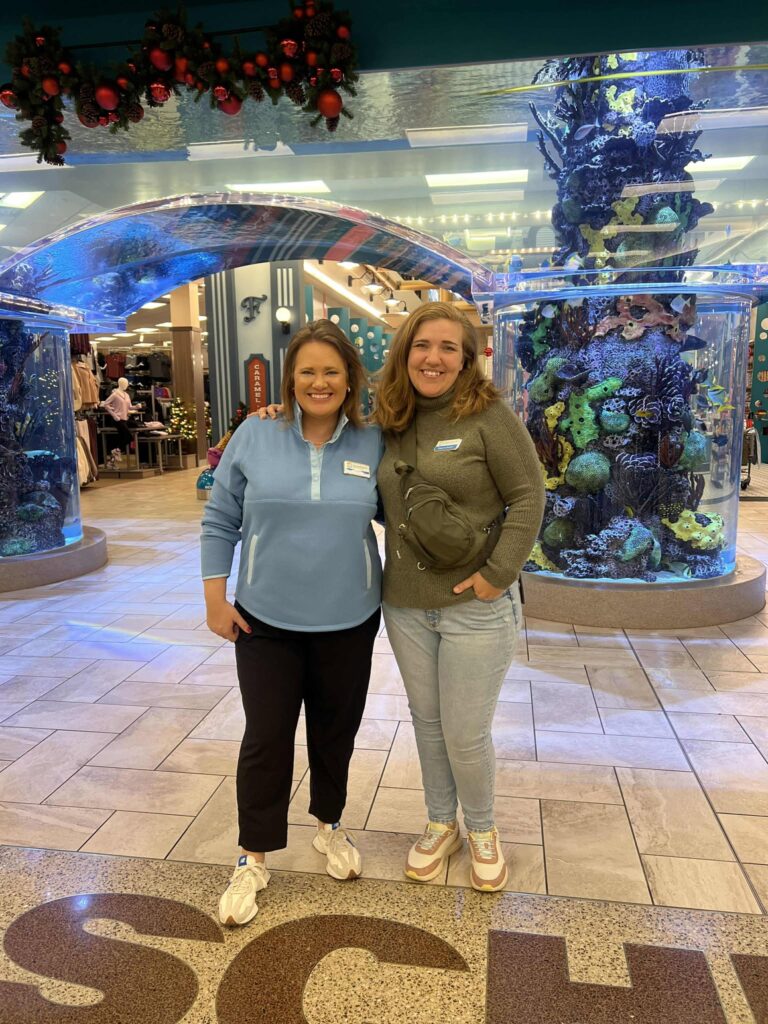 Two people are standing and smiling in front of an aquarium display inside a shopping mall. One person is wearing a light blue top, and the other is in a gray sweater. The floor is tiled, and there are holiday decorations above.