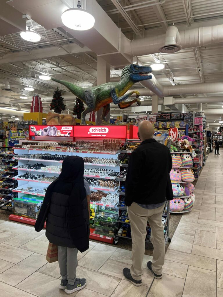 A child and an adult stand in a store aisle looking at a large model dinosaur displayed above shelves filled with toys. The aisle is brightly lit and various plush toys are visible to the right.