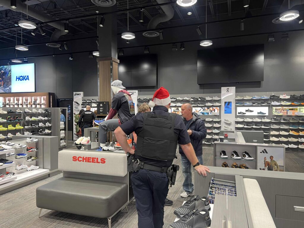 Two uniformed individuals and a woman interact with a person wearing a gray hoodie and hat sitting on a bench in a shoe store. Shelves display various athletic shoes and a large screen shows a brand logo in the background.