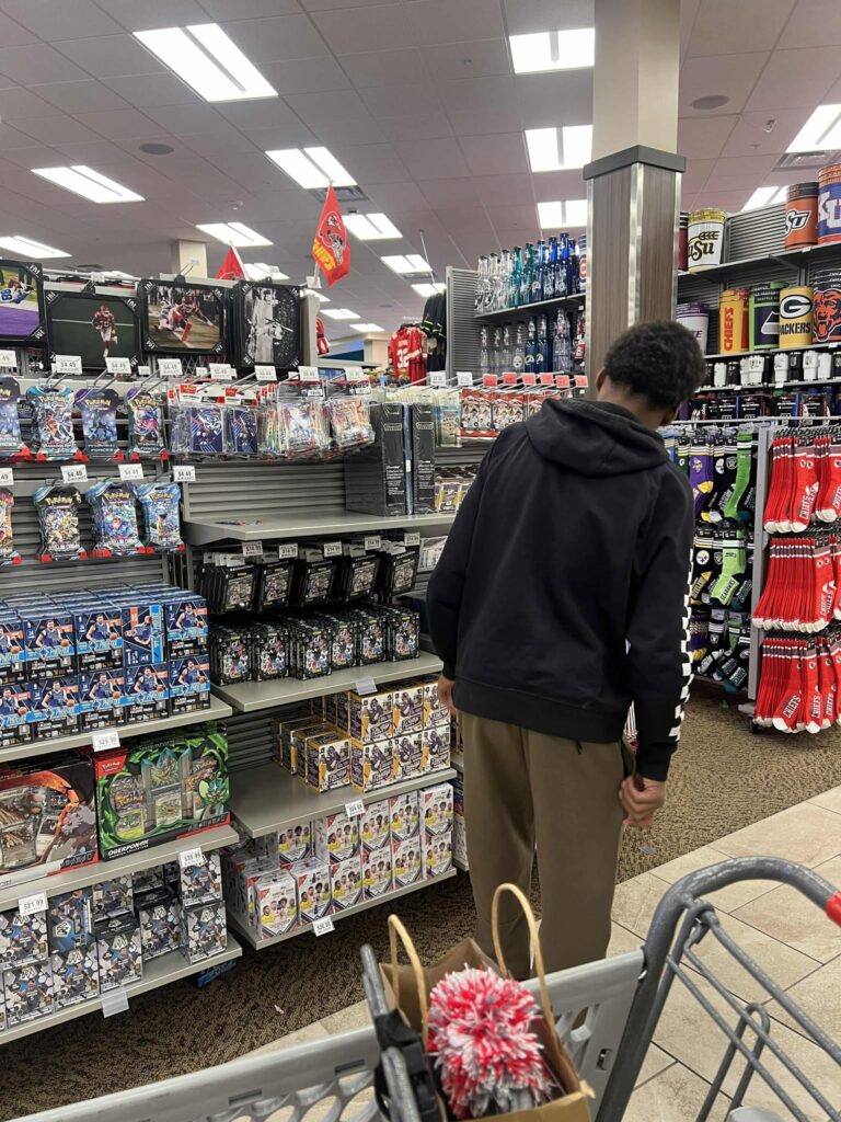 A person wearing a black hoodie and beige pants is standing in front of a shelf stocked with collectible toys and figures in a store. A shopping cart with items is partially visible in the foreground.