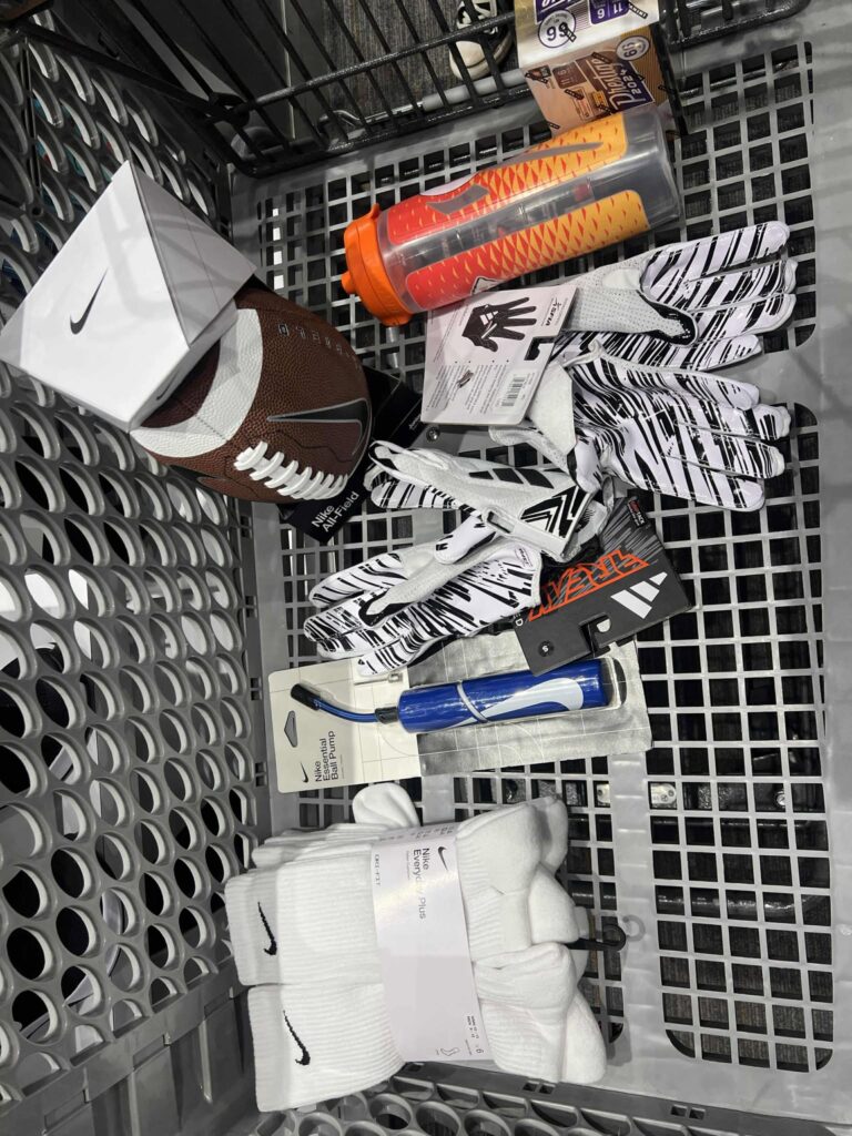 A shopping cart contains a Nike football, two pairs of patterned sports gloves, a pack of socks, an orange and yellow shaker bottle, a tube of ChapStick, and a box of Swiss Miss hot chocolate mix.