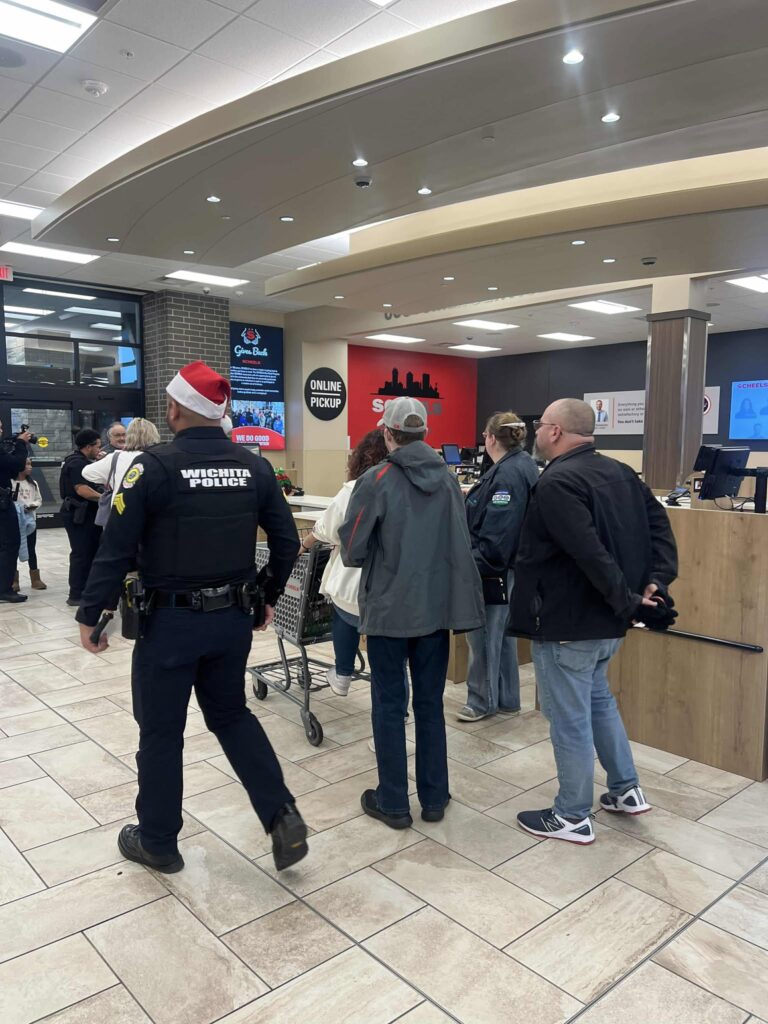 A police officer stands in a store wearing a Santa hat and uniform, talking to a group of people near the online pickup counter. The store has tiled flooring and bright overhead lighting. Customers and staff are in the background.