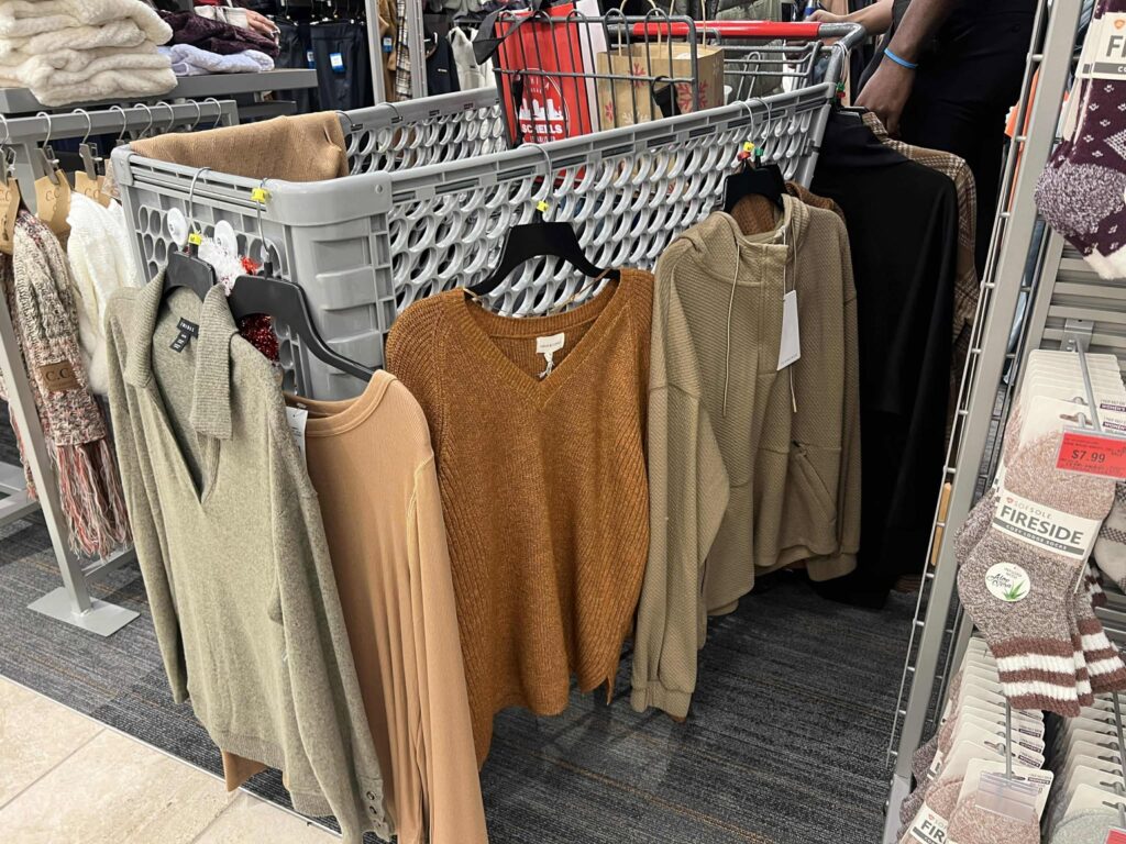 A shopping cart is filled with various sweaters in neutral colors like brown, beige, and gray. They are displayed on hangers and placed along a store aisle. Nearby shelves hold folded clothes and a pair of patterned socks is visible in the foreground.