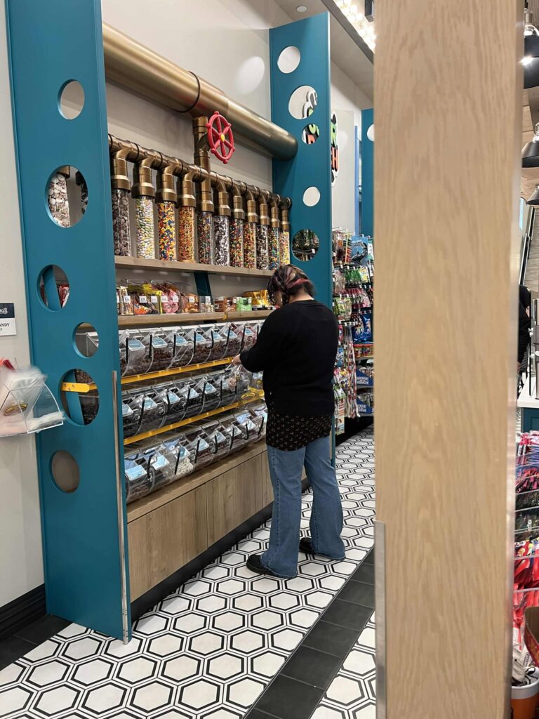 A person is standing in front of a large candy display in a store. The display features clear tubes filled with various colorful candies and bags of candy on shelves below. The floor has a black and white hexagonal tile pattern.