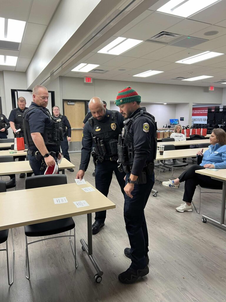 Police officers stand around a table in a room with fluorescent lighting. One officer is wearing a colorful knit hat. A few people are sitting at nearby tables, and papers are spread on the table. The setting appears to be a break room or meeting area.