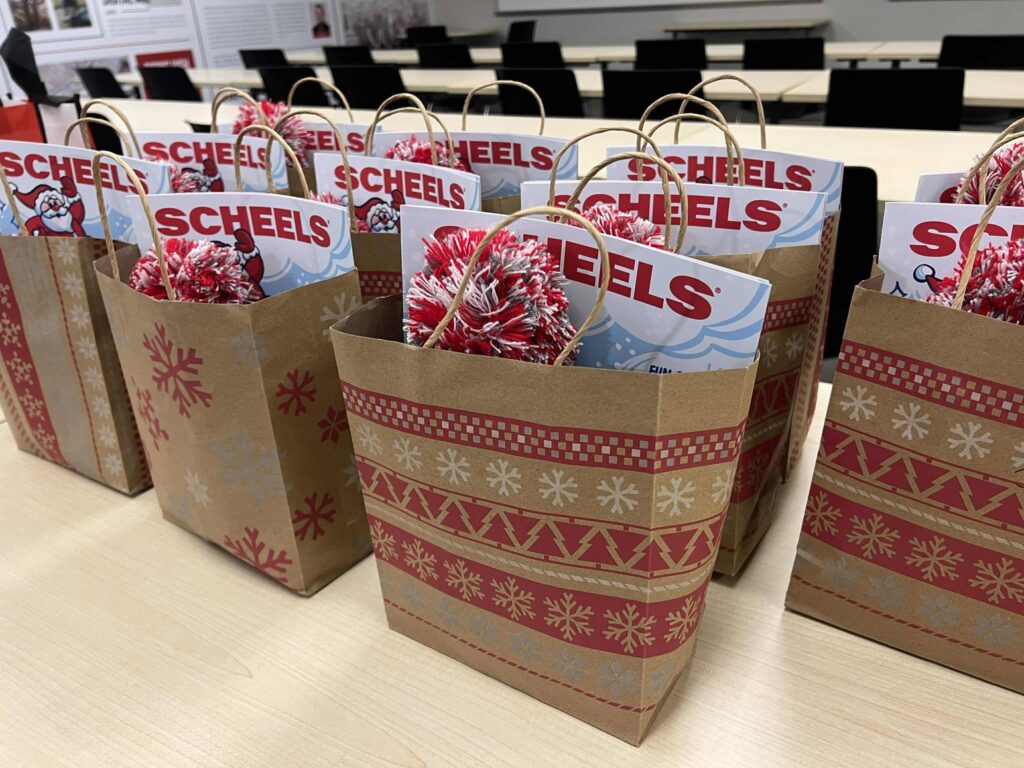 Gift bags are lined up on a table, each containing a Scheels store catalog or flyer. The bags are decorated with red and white holiday patterns featuring snowflakes and festive designs. The background shows tables and chairs.