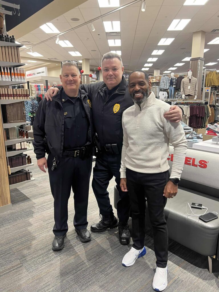 Three men are standing together inside a store. Two are wearing police uniforms, and one is in a light-colored sweater and black pants. They are smiling and standing near shelves and a bench with clothes in the background.