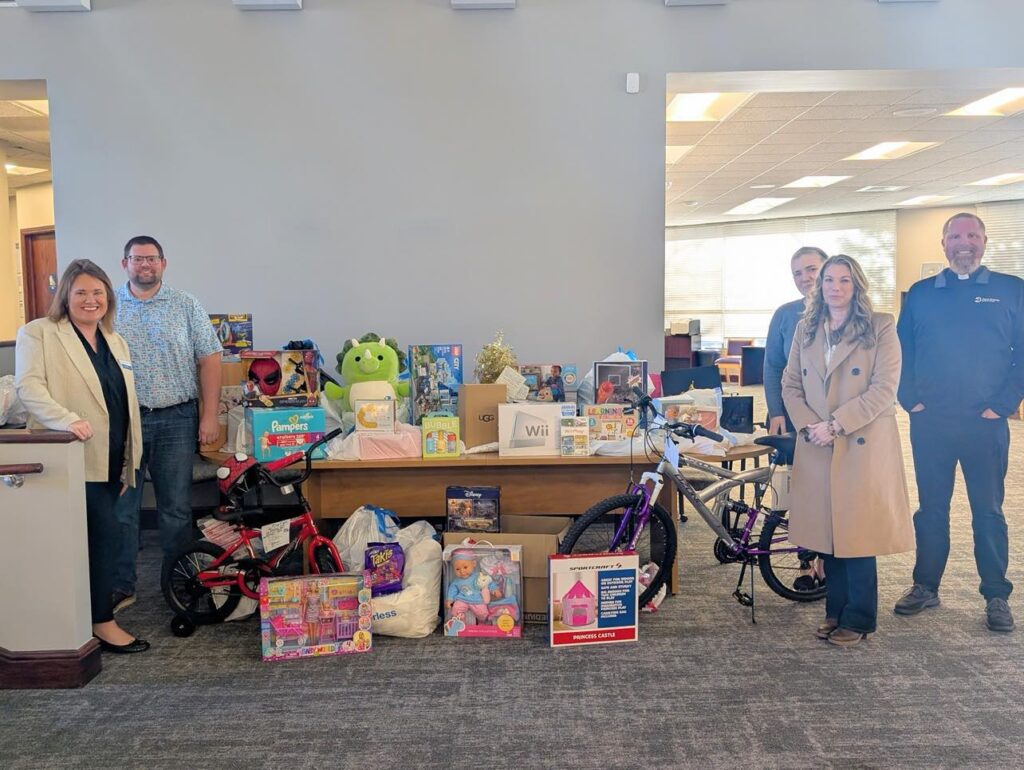 Five people stand in a room next to a table filled with various donated items, including toys, bicycles, and games. The setting appears to be an office space with gray carpeting and a large window in the background.