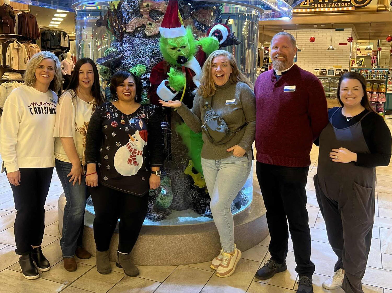 Six people stand in a shopping area in front of a decorative tank with a large, green figure dressed in a Santa hat. Some are wearing festive sweaters. The scene is bustling with holiday decorations.