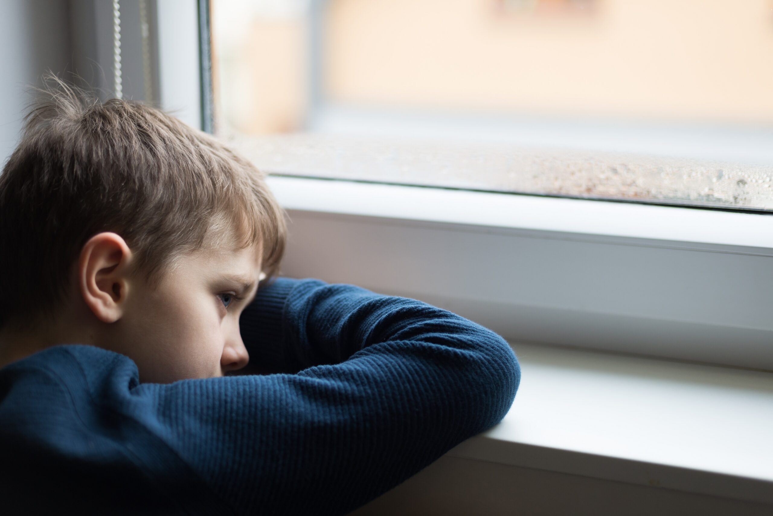 A young boy with short brown hair rests his arms and chin on a windowsill, gazing outside. He is wearing a dark blue long-sleeved shirt. The window has small raindrops on the glass, suggesting it is raining outside.