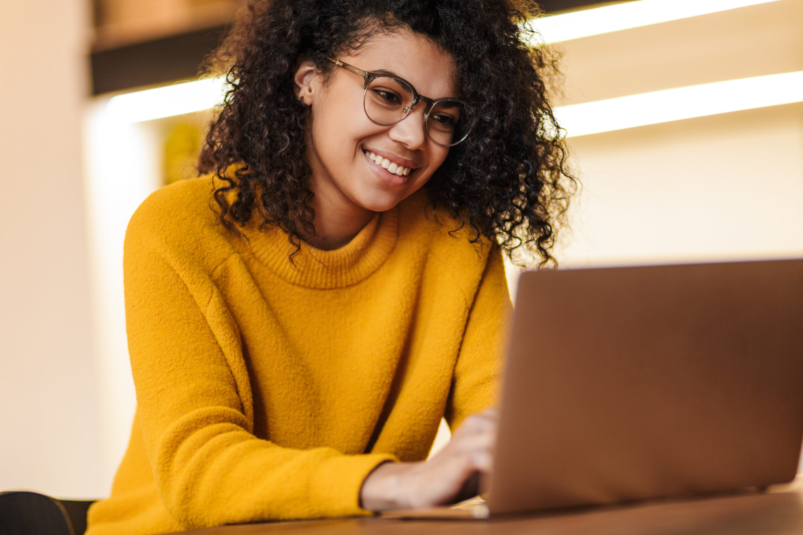 A person wearing glasses and a yellow sweater is smiling while typing on a laptop. The background includes soft-focus lighting and warm-toned decor.