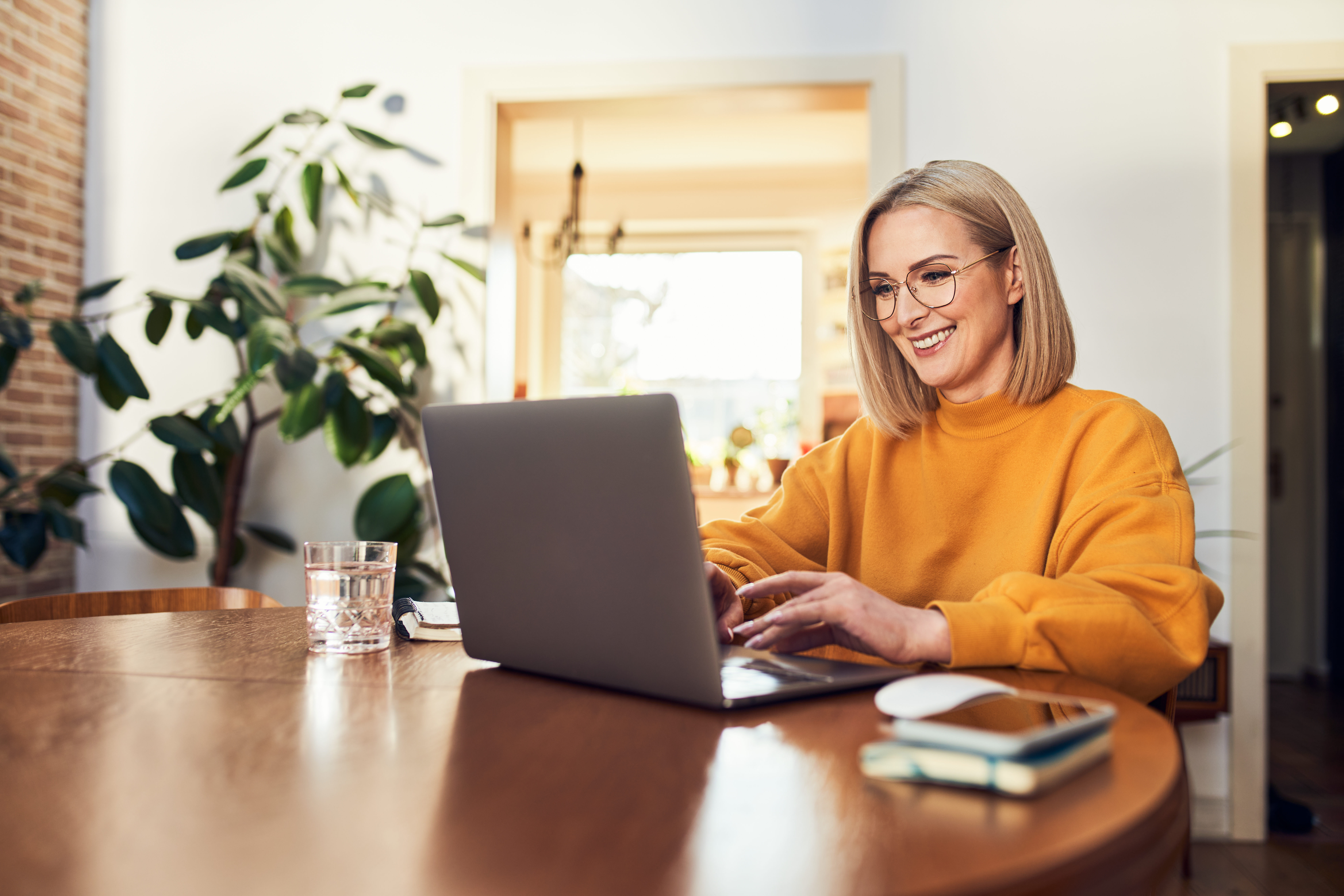 A woman with blonde hair and glasses is sitting at a wooden table, using a laptop. She is wearing a yellow sweater and smiling. On the table are a glass of water, a notebook, and a computer mouse. A large plant is in the background.