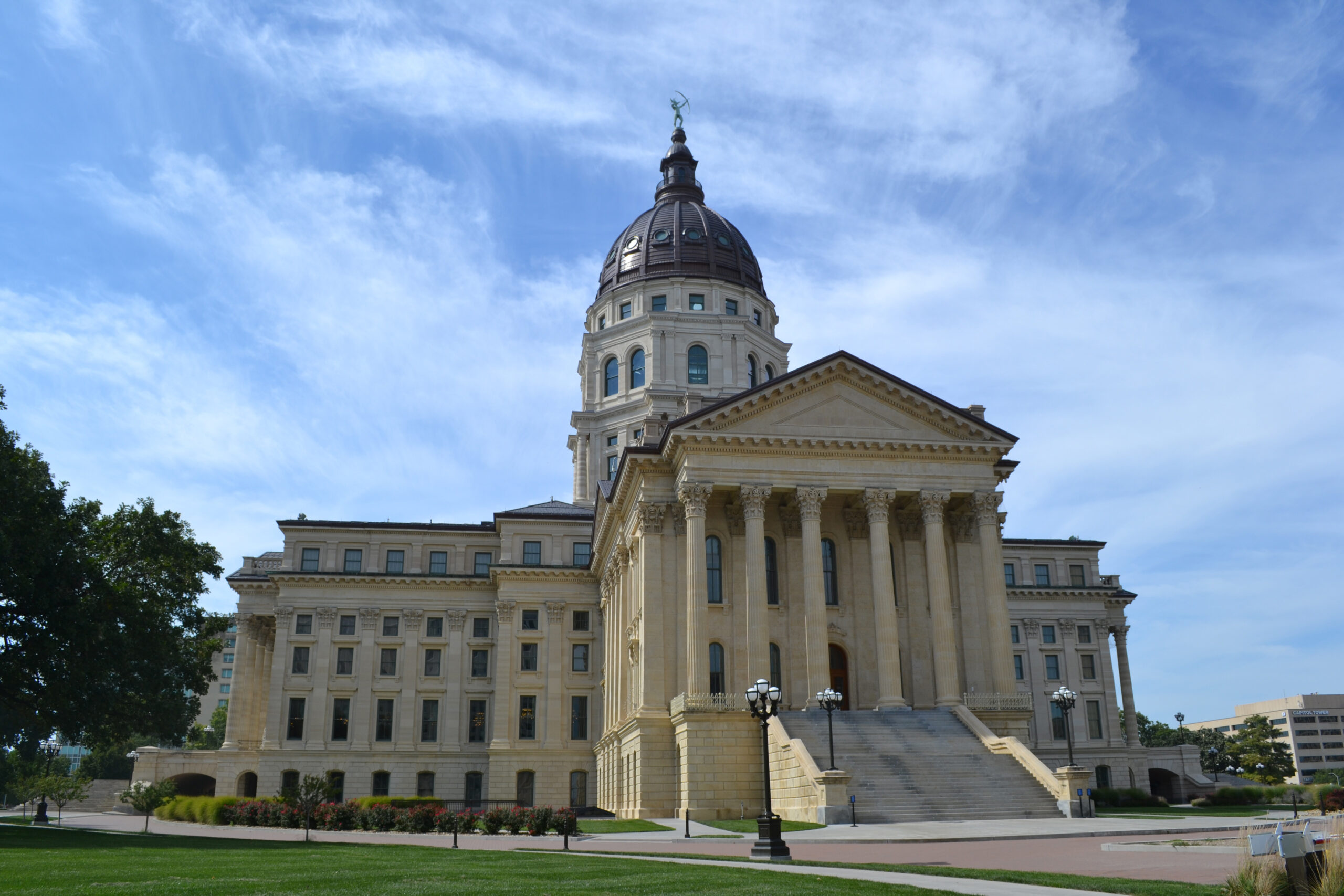 The image shows a large, historic government building with a domed roof, tall columns, and steps leading up to the entrance. The sky is partly cloudy, and there are well-maintained lawns and trees surrounding the building.