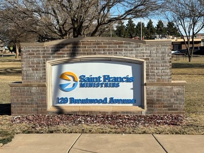 A brick sign with "Saint Francis Ministries" written in white and blue letters, featuring a circular emblem to the left. Below the logo is the address "139 Brentwood Avenue." Trees and grass are visible in the background.