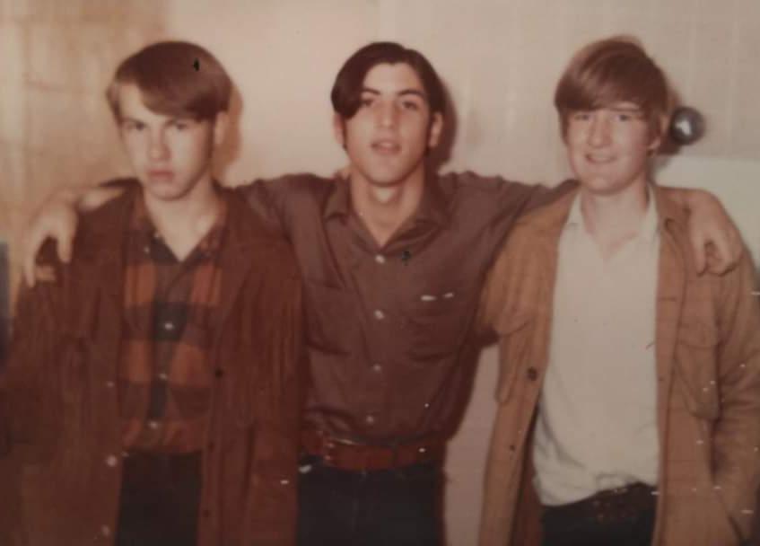 Three young men stand together indoors, smiling and with arms around each other. They are wearing casual shirts and jackets. The background features a light-colored wall.