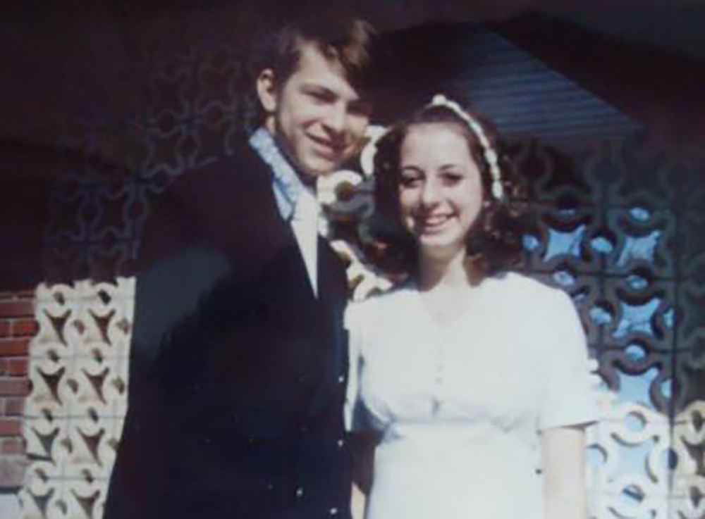 A young couple stands outside in front of a decorative wall. The man is wearing a dark suit and the woman is in a white dress with a headband. Both are smiling.