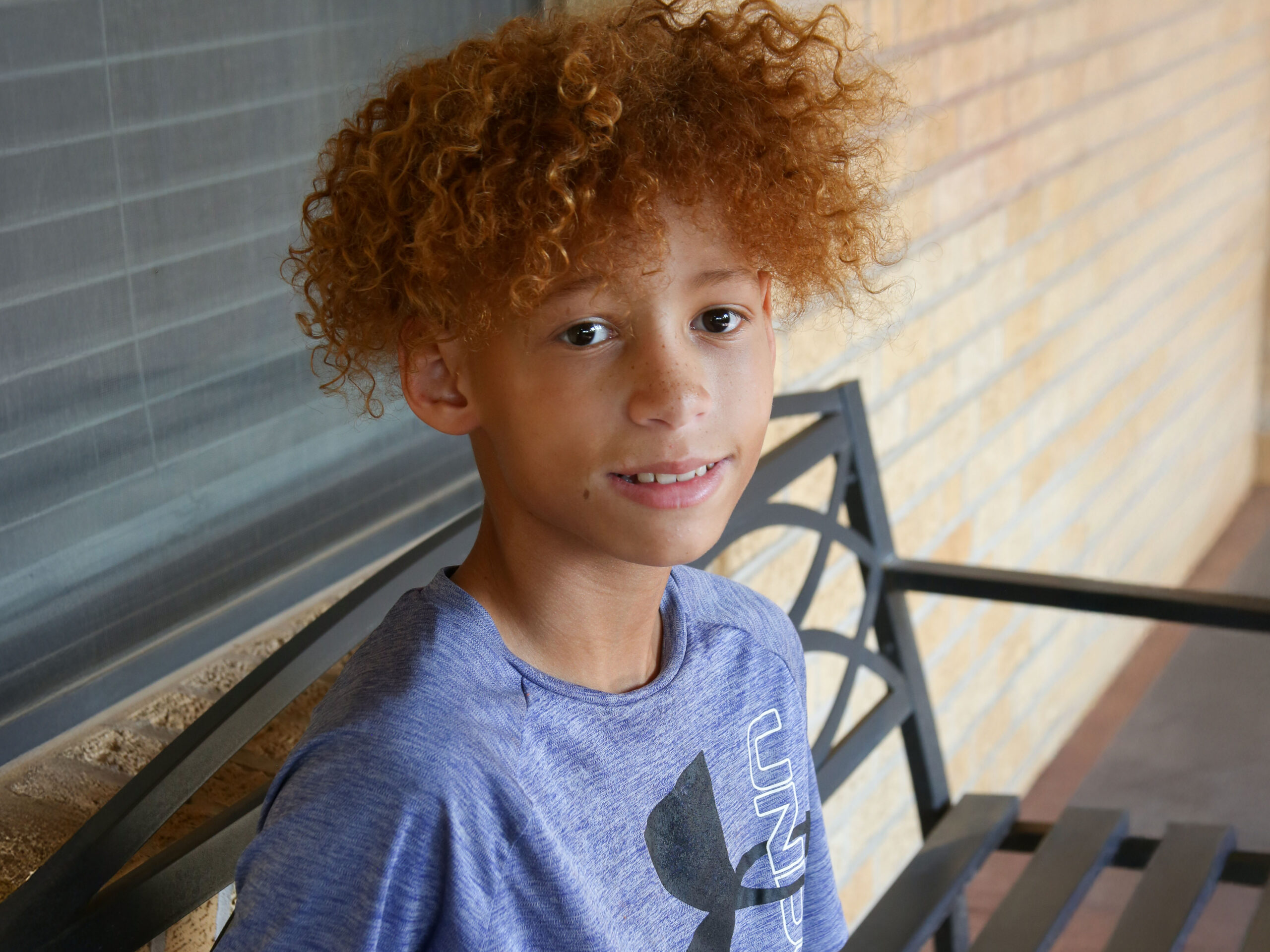 A young person with curly, light brown hair sits on a metal bench. They are wearing a gray T-shirt and are looking at the camera with a slight smile. The background features a brick wall.