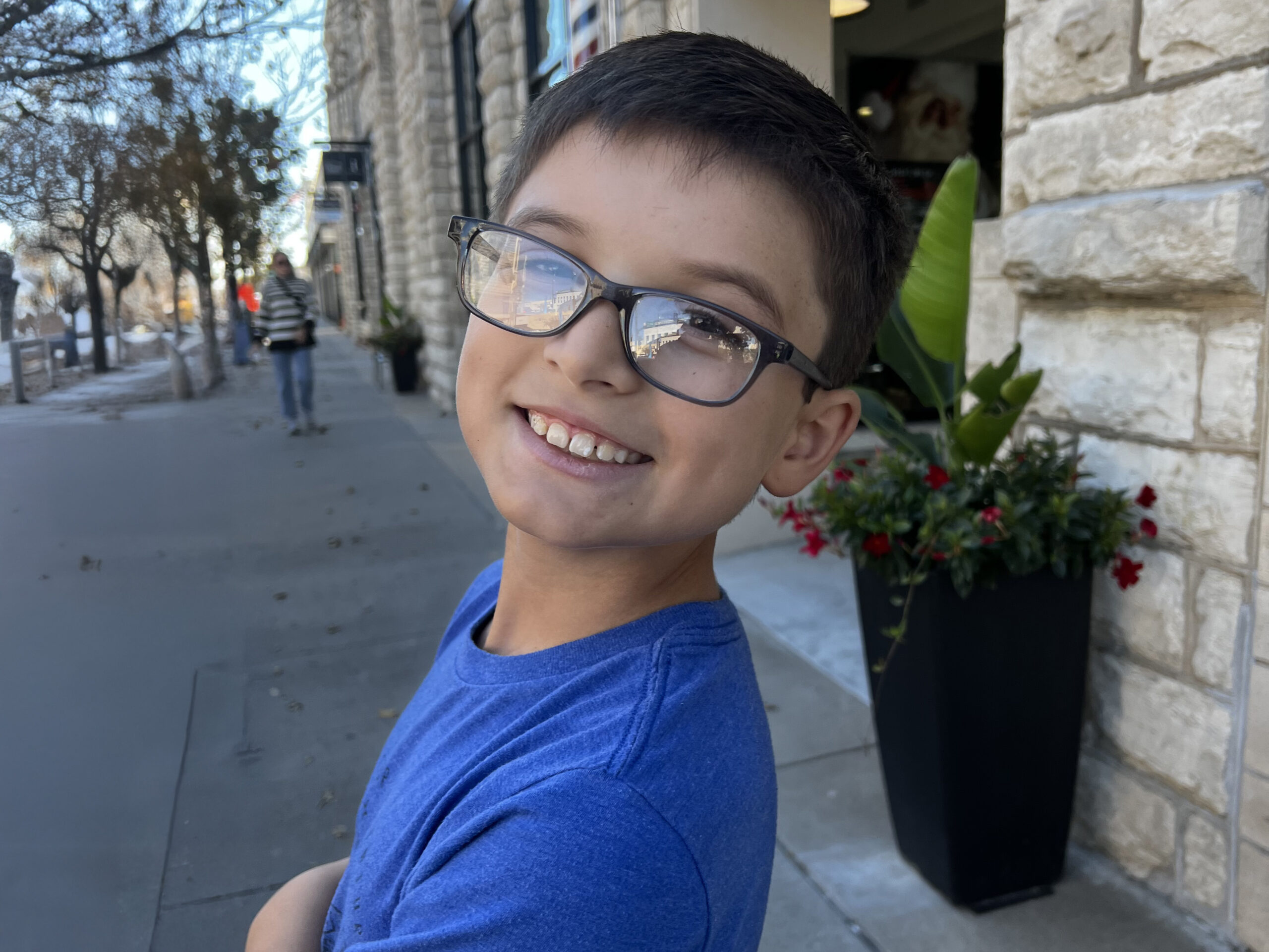 A young boy with glasses and a blue shirt is smiling at the camera while standing on a sidewalk. There are stone buildings and a large plant in the background, and a person walking away in the distance.