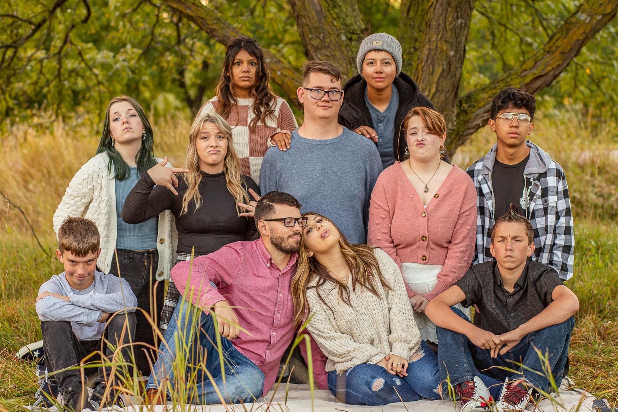 A group of 12 people pose outdoors on a grassy area with trees in the background. They display various expressions and poses, some sitting and others standing. The mood is casual and relaxed.