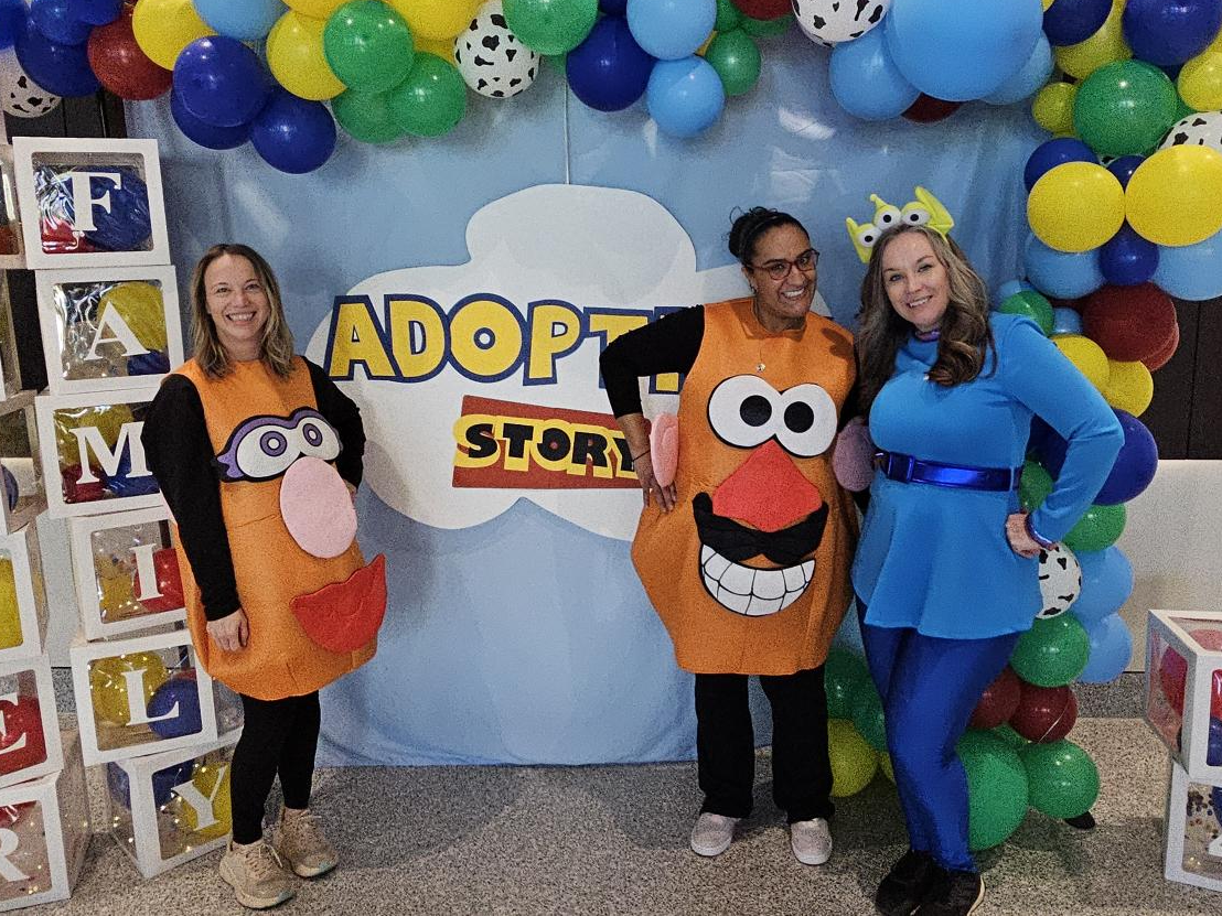 Three people are wearing costumes, two as Mr. Potato Head and one in a blue outfit and hat with antennae. They stand in front of a backdrop that reads "Adoption Story," surrounded by colorful balloons and blocks spelling "FAMILY.