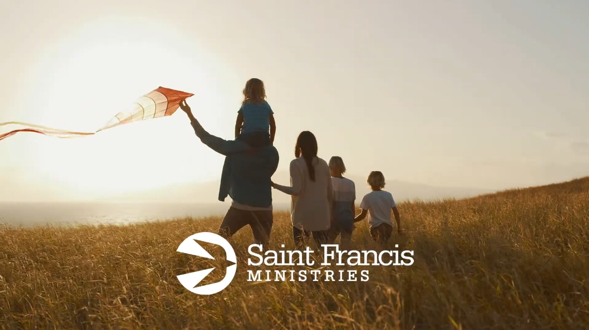 A family of four walks through a grassy field at sunset. One person carries a child on their shoulders, while another holds a kite. The Saint Francis Ministries logo is displayed in the foreground.