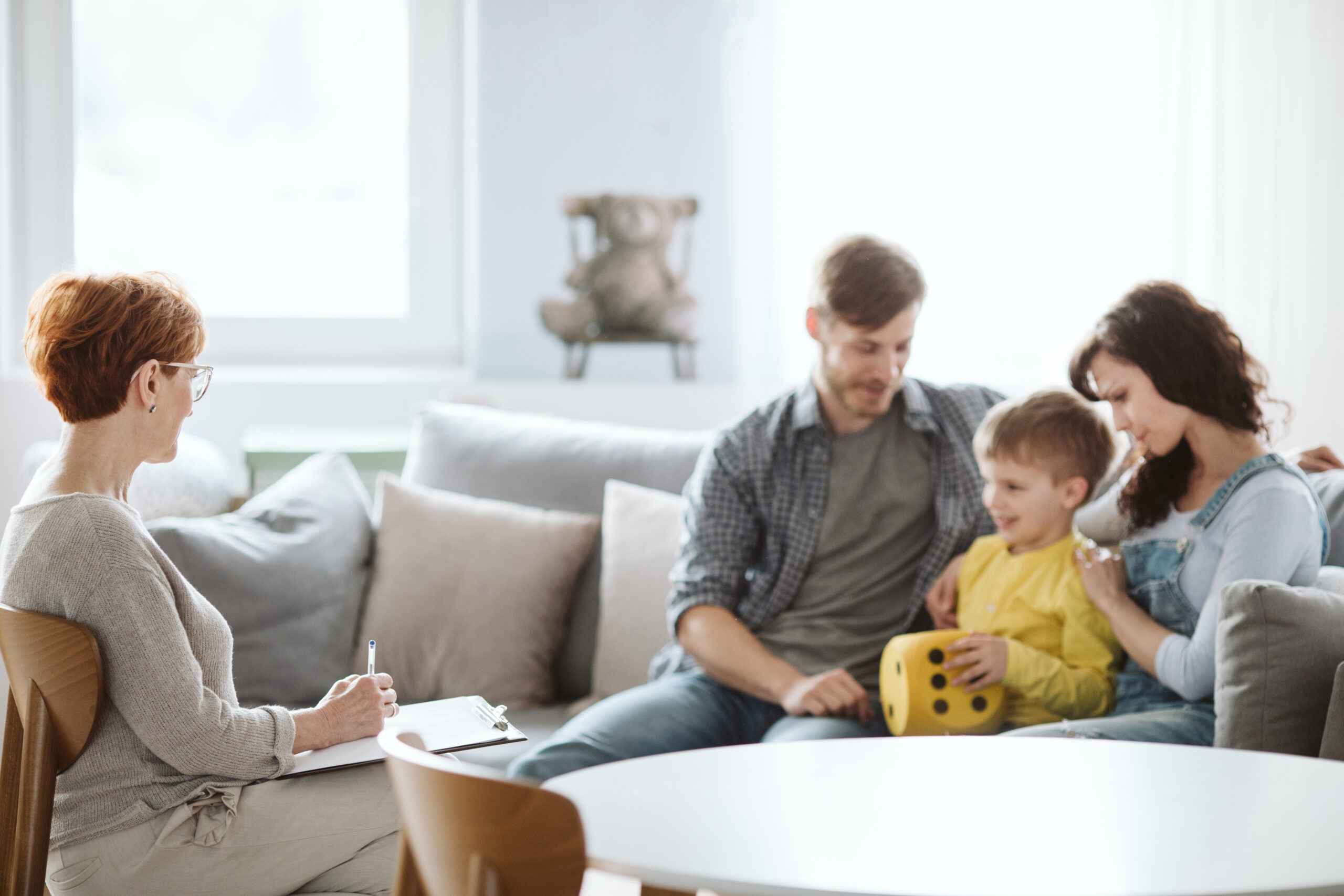 A family sits on a sofa with a child holding a large yellow die. A man and woman sit on either side of the child, while another woman with short hair and glasses sits nearby, writing on a clipboard. The room is softly lit and furnished with light colors.