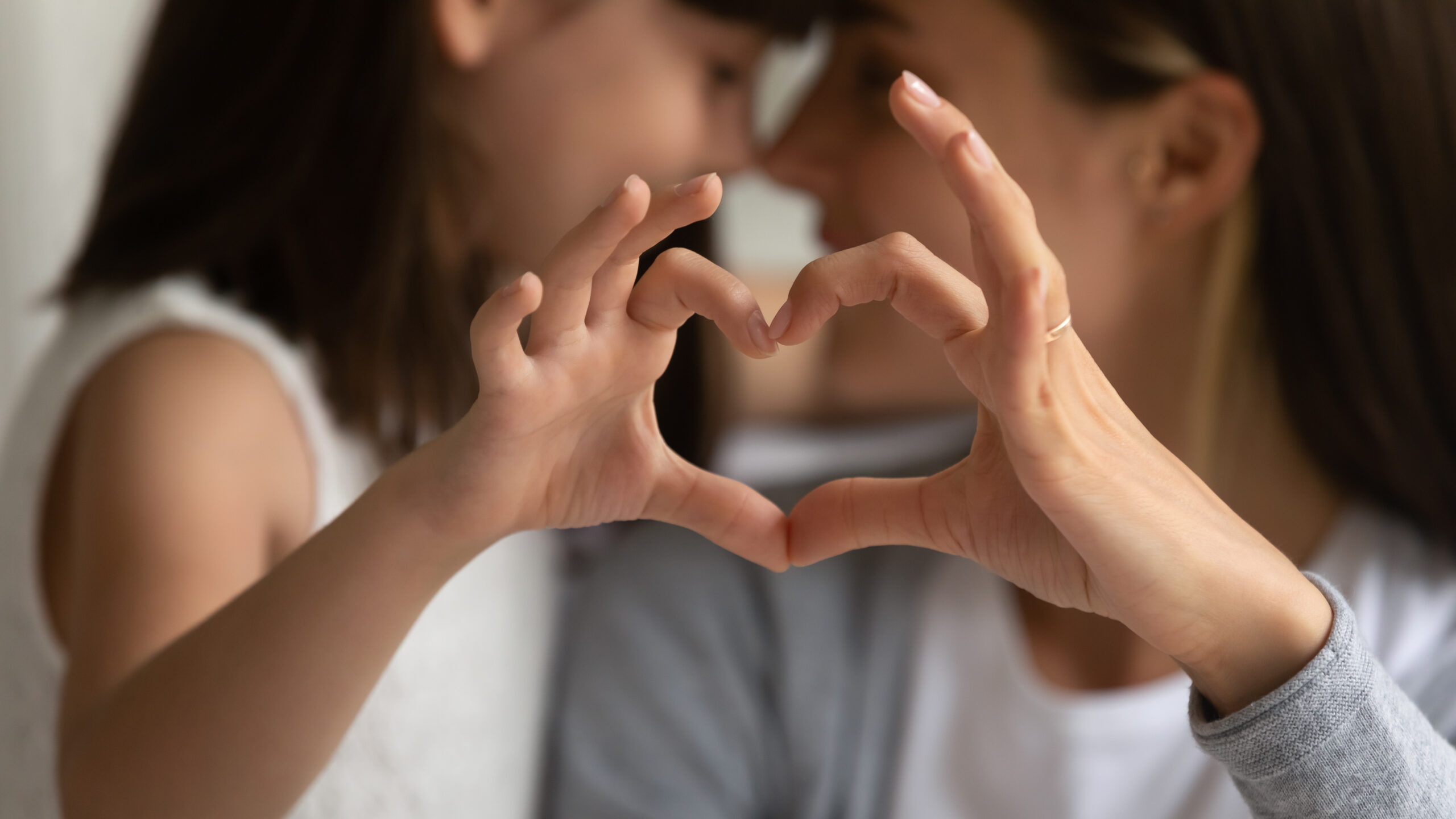 Two people, an adult and a child, form a heart shape with their hands in the foreground, their faces close together in the background. The focus is on their hands, showing a sense of affection and connection.