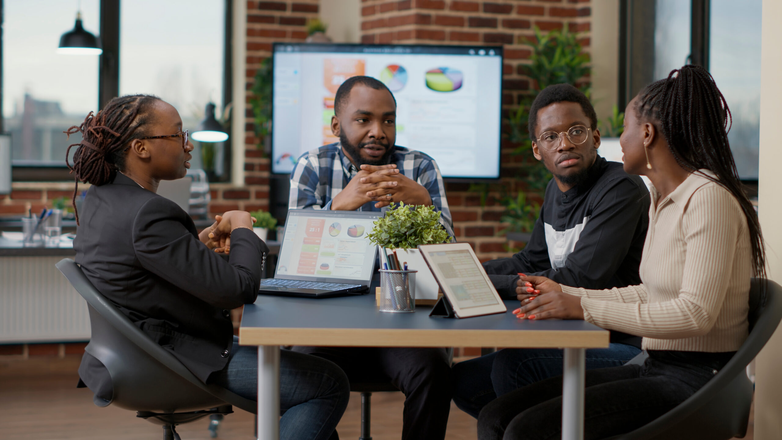 Four people sit at a table in an office setting, engaged in discussion. Two people use laptops, while a digital presentation is displayed on a screen in the background. A small potted plant is on the table.