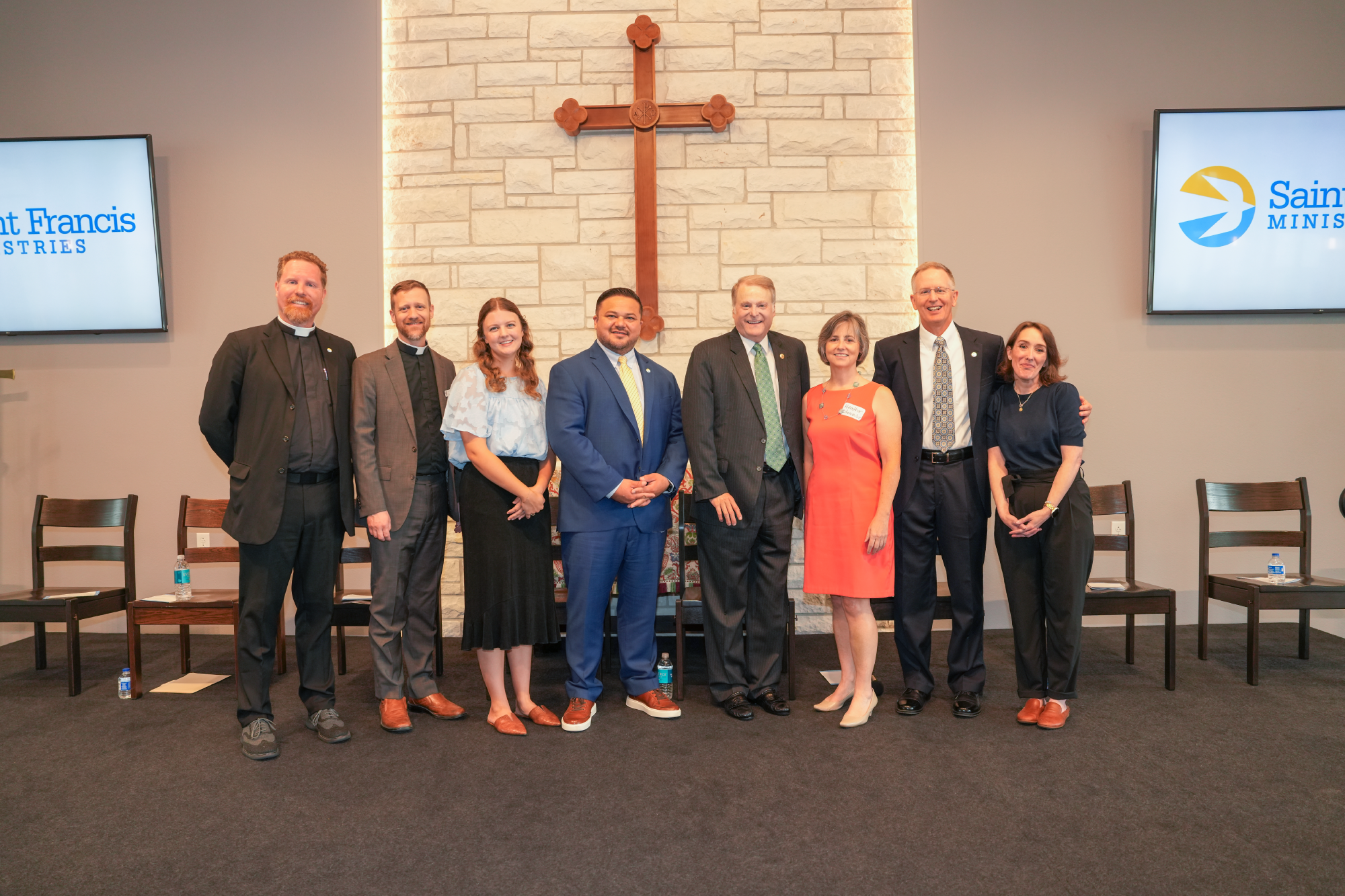 A group of nine people standing in a row on a stage, dressed in formal attire. They are in front of a stone wall with a large wooden cross in the center. Two screens on either side display "Saint Francis Ministries.