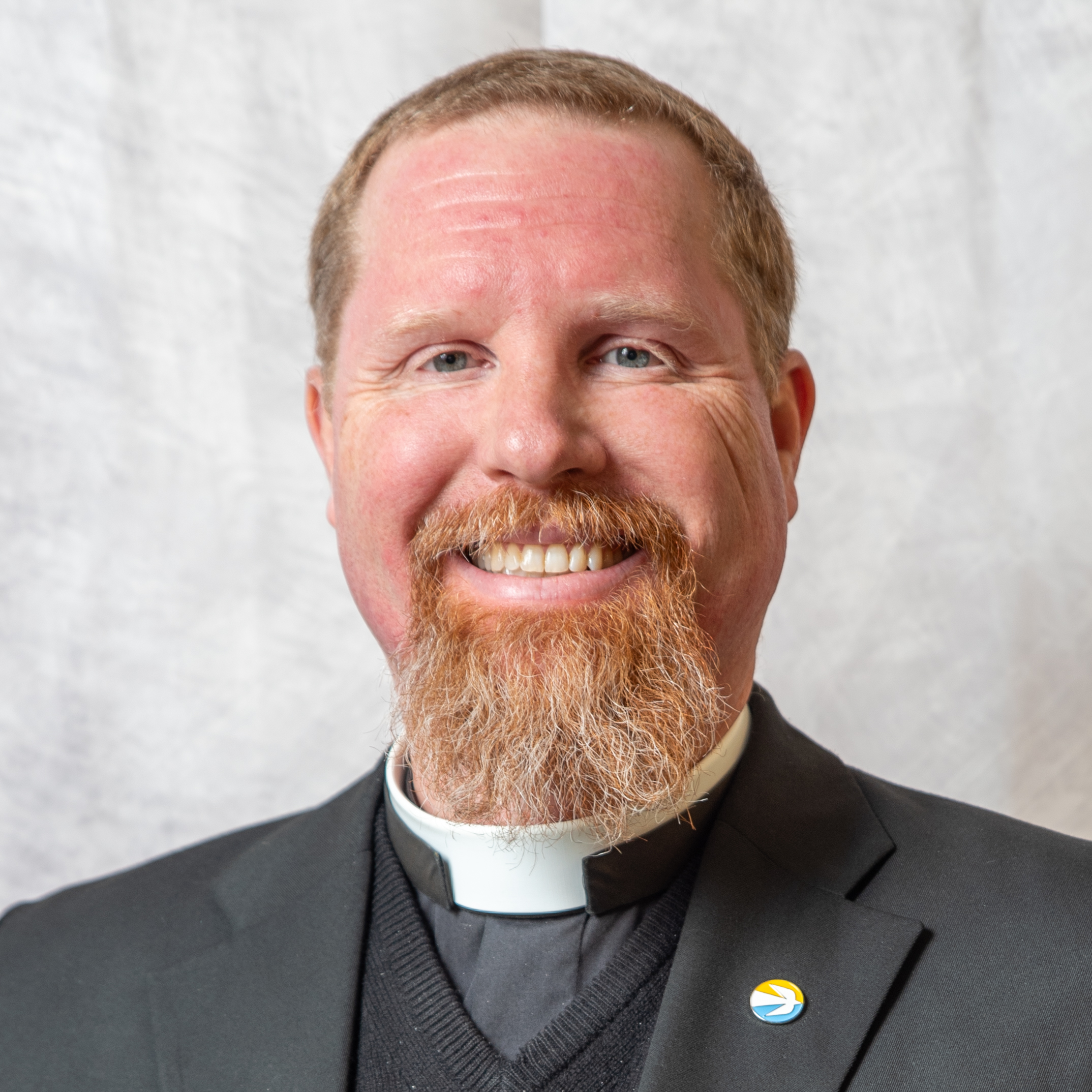 A man with a reddish beard and short hair is wearing a black suit with a clerical collar and a pin on the lapel. He is smiling and standing in front of a light, textured background.