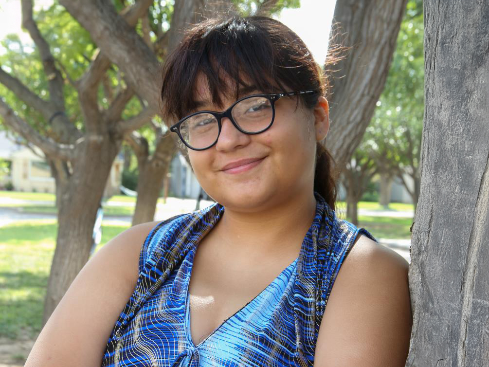 A person with glasses and bangs is smiling, leaning against a tree in a park. They are wearing a blue patterned sleeveless top. Trees and green foliage are in the background.