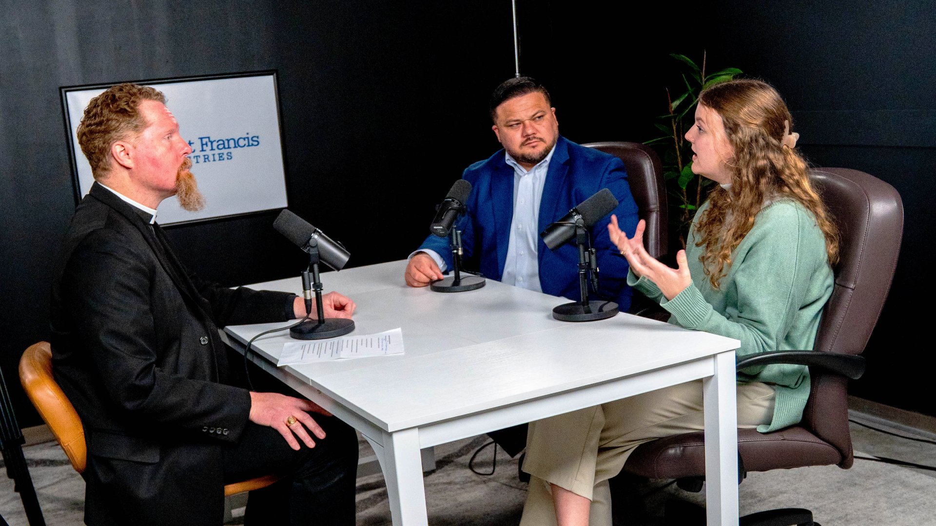 Three people are seated around a white table with microphones in front of them, appearing to record a podcast or interview. The setup is in a studio with a dark background and a sign reading "Francis" on the wall.