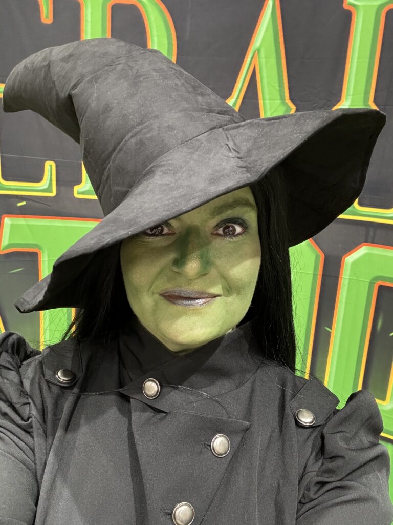 A person dressed as a witch with green face paint, wearing a black pointed hat and a black outfit with decorative buttons. The background features large green and yellow letters on a black surface.