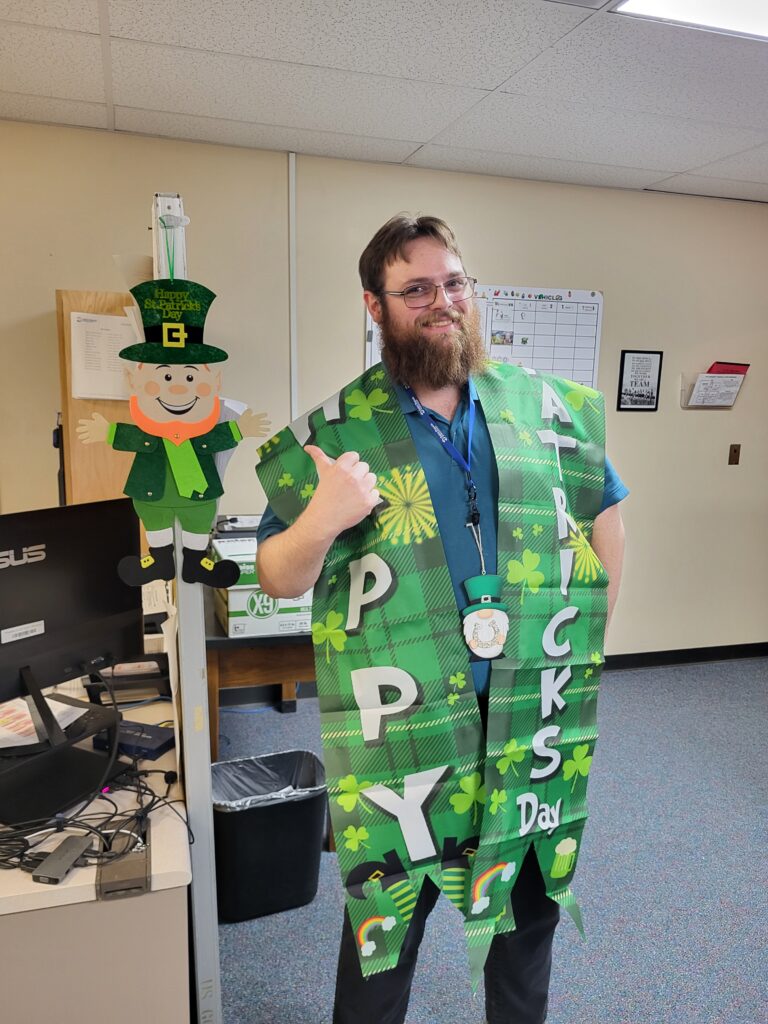A man with a beard stands in an office, wearing a green St. Patrick's Day-themed outfit. The outfit has clover patterns and text reading "Happy St. Patrick's Day." He is giving a thumbs-up. A leprechaun decoration is visible in the background.
