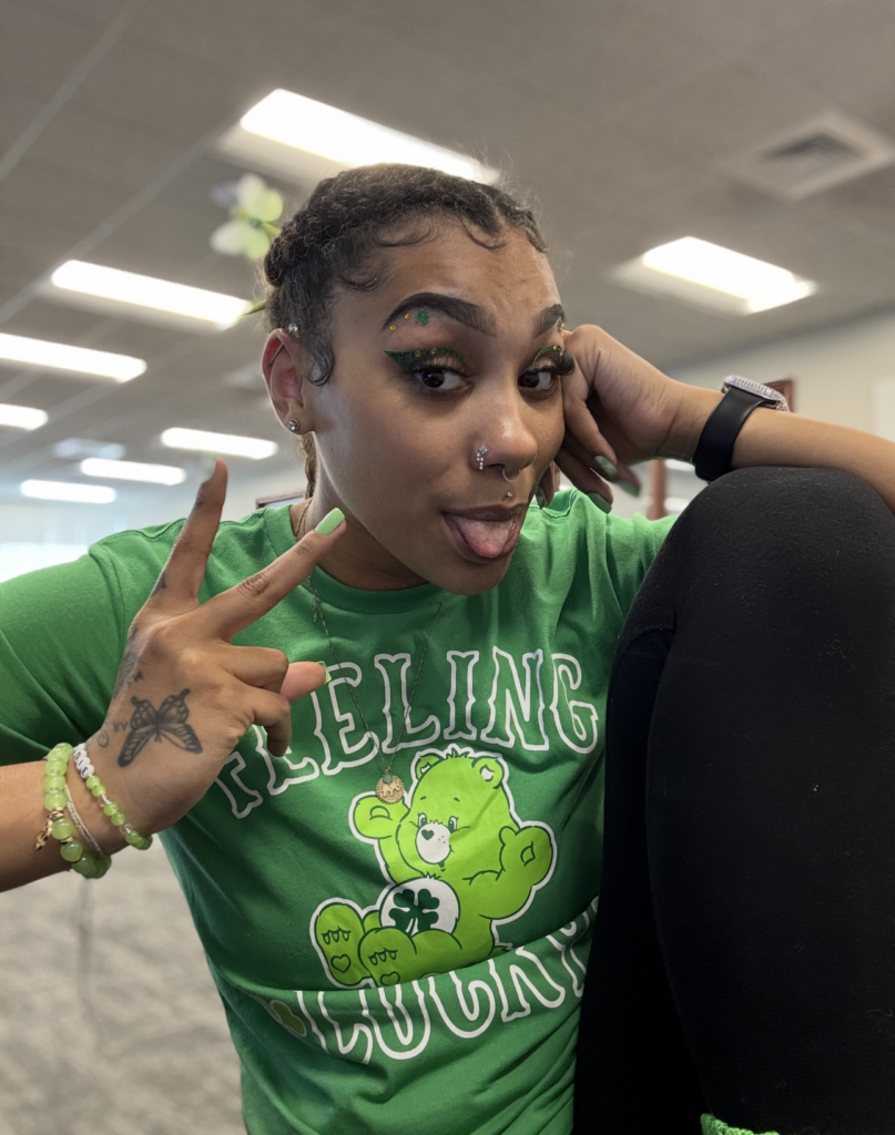 A person wearing green eyeshadow and a green T-shirt with a bear design smiles and gestures with their hand. They have piercings and a tattoo on their hand. The background is an indoor space with a ceiling and lights.