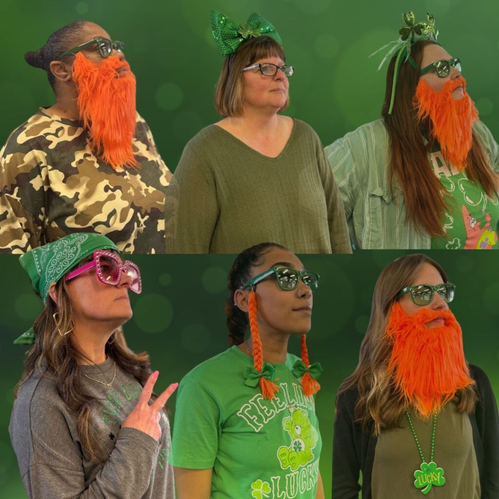 A group of people wearing green-themed attire, with some sporting fake orange beards and sunglasses. They are in front of a green background. One person wears a green bow, another has heart-shaped glasses, and a "Lucky" necklace is visible.