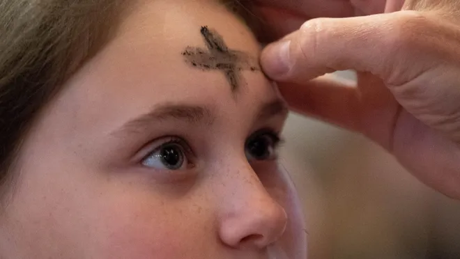 A person is having a cross made of ash applied to their forehead by another person's hand. The background is blurred, focusing on the face and the hand with ash.