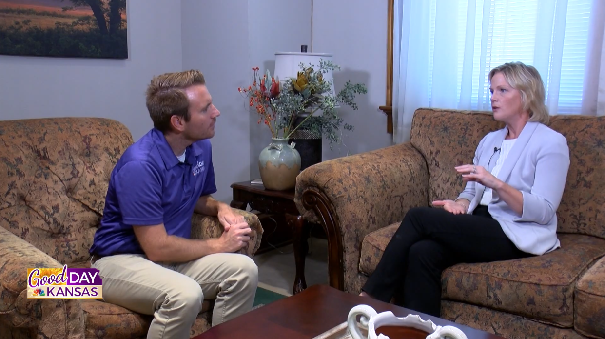 A man and a woman are seated on patterned sofas in a living room setting, engaged in conversation. A table with decorative items is between them. The "Good Day Kansas" logo is in the bottom left corner.