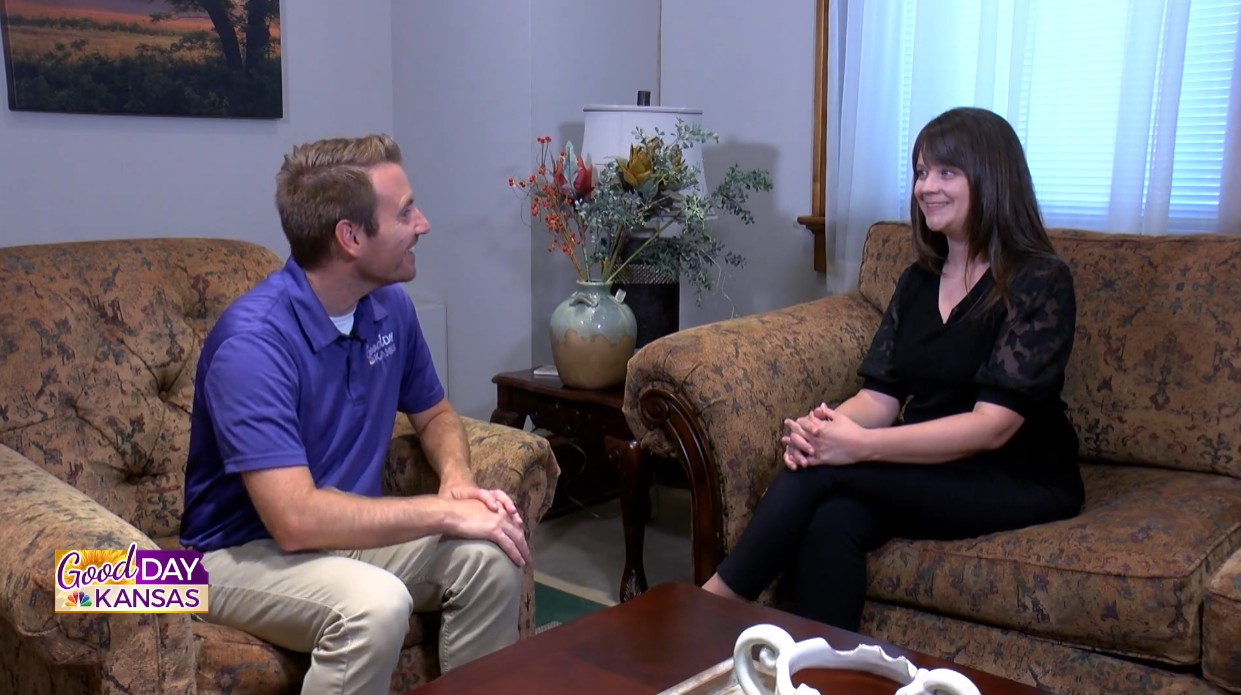 A man in a purple polo shirt sits on a couch talking to a woman in a black blouse seated on another couch. The setting is a living room with a vase of flowers on a side table and the "Good Day Kansas" logo in the corner.