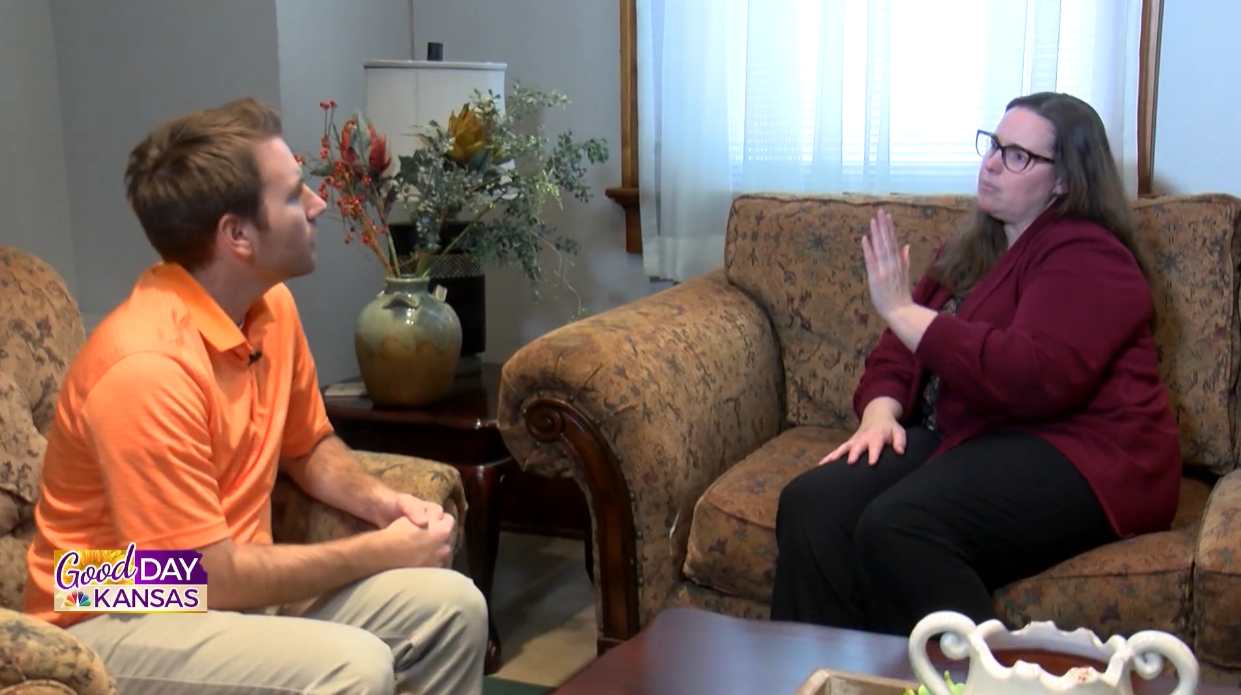 A man and a woman are sitting on a floral-patterned couch talking. The woman is gesturing with her hand. They are in a living room with curtains, a large vase with flowers on a side table, and "Good Day Kansas" is visible on the screen.