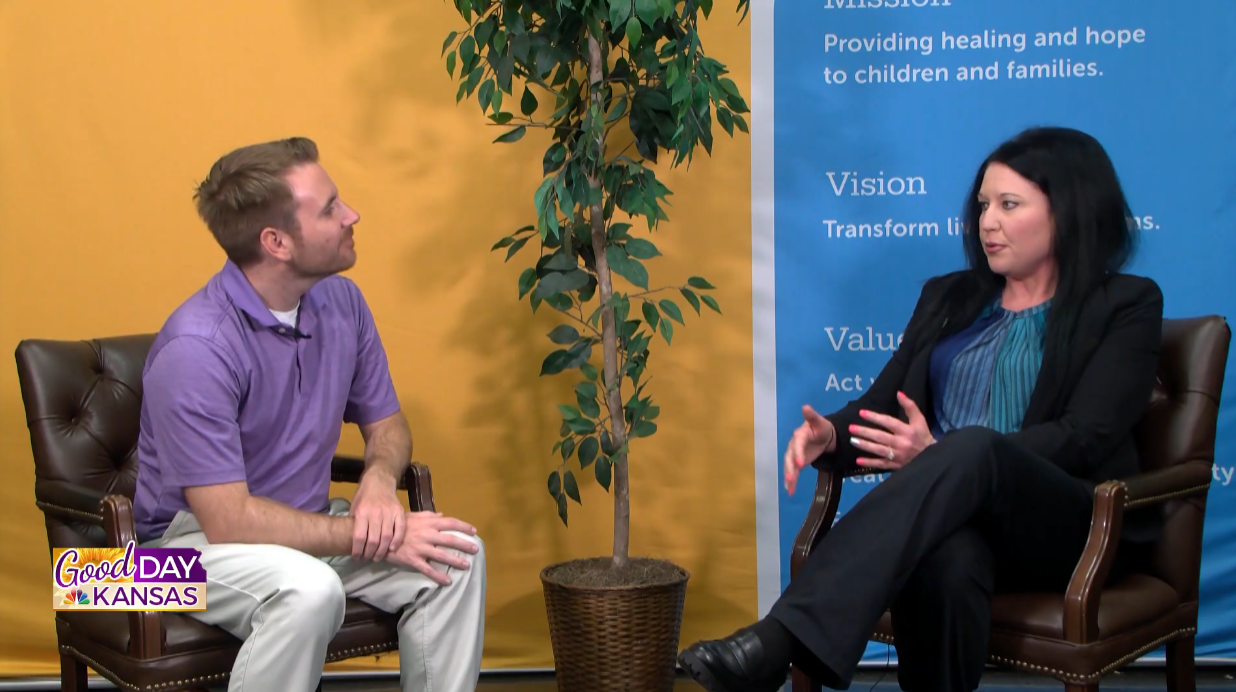 A man and a woman sit in chairs near a potted plant, engaged in conversation. The backdrop features text about mission, vision, and values. The image is labeled "Good Day Kansas.