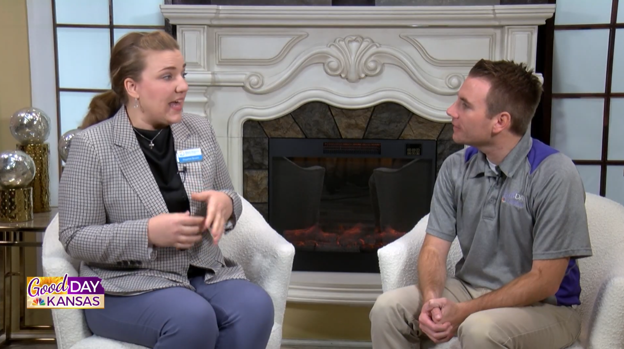 Two individuals are seated and engaged in conversation in a studio setting. The person on the left gestures with hands while speaking. They sit in front of an ornate fireplace. A "Good Day Kansas" graphic is visible in the bottom left corner.