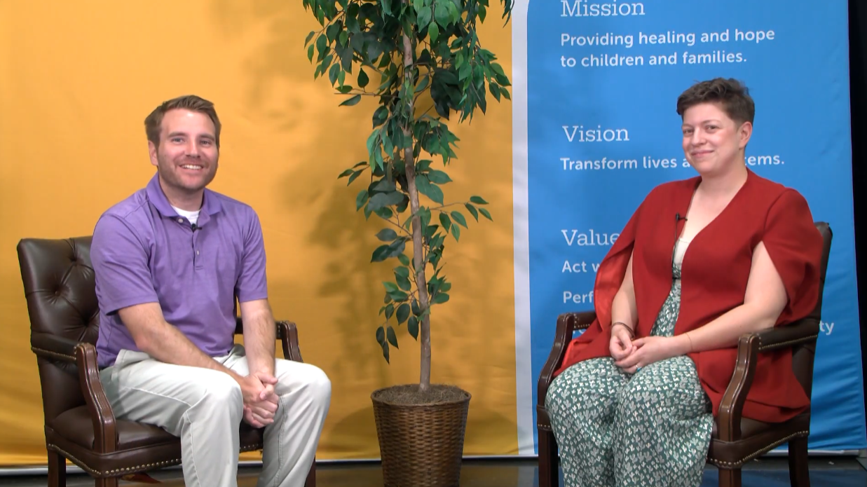 Two individuals sit on leather chairs with a potted tree between them. The backdrop includes a yellow and blue screen with text outlining an organization's mission, vision, and values. The person on the left wears a purple shirt; the person on the right wears a red top.