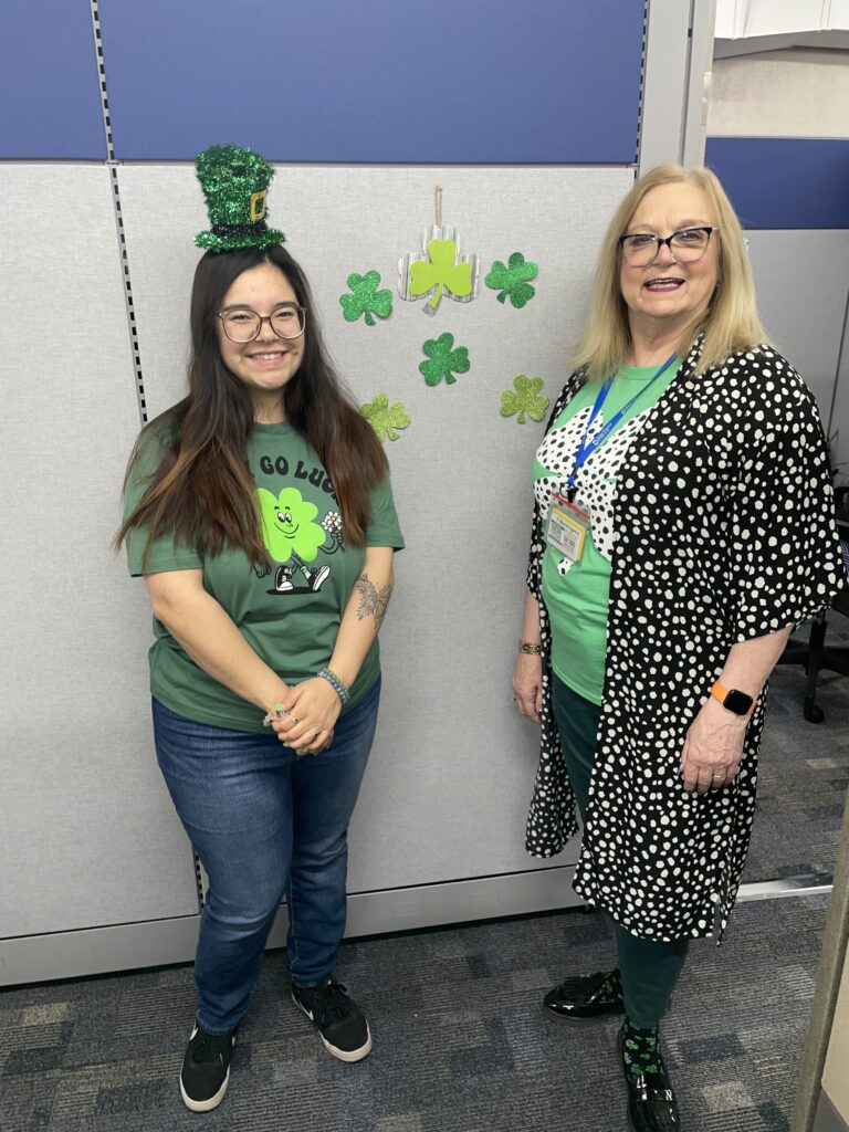 Two women stand in front of a gray partition decorated with shamrocks. The woman on the left wears a leprechaun hat and a green shirt. The woman on the right wears a polka dot cardigan with a green top. Both are smiling and posing for the camera.