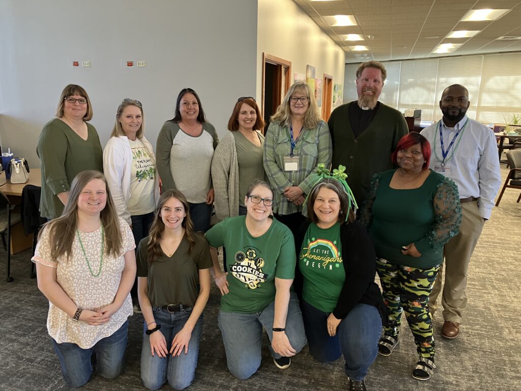 A group of thirteen adults standing and sitting indoors. Most are wearing green-themed clothing, with some wearing shirts with St. Patrick's Day designs. They are in an office setting with desks and chairs in the background.