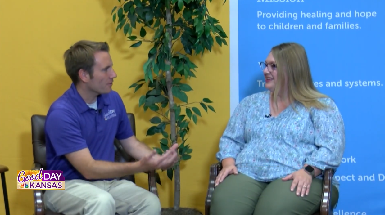A man and a woman sit facing each other on a set. The man wears a purple polo shirt and beige pants, gesturing while speaking. The woman wears glasses, a floral blouse, and green pants, smiling and listening. A plant and signage are in the background.