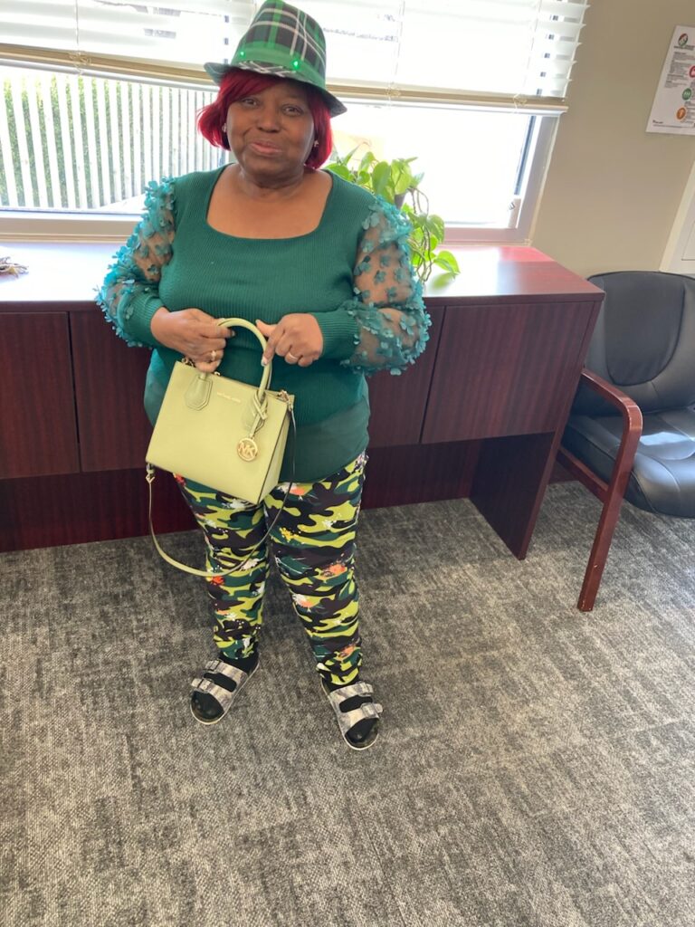A person wearing a green blouse with sheer, patterned sleeves, camouflage pants, and a green hat stands in an office. They are holding a light green handbag and wearing black sandals. A plant and a chair are in the background.